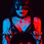 An image of a woman in lingerie with leather bondage straps. She is holding a riding crop, and starting BDSM play as she seductively looks into the camera.