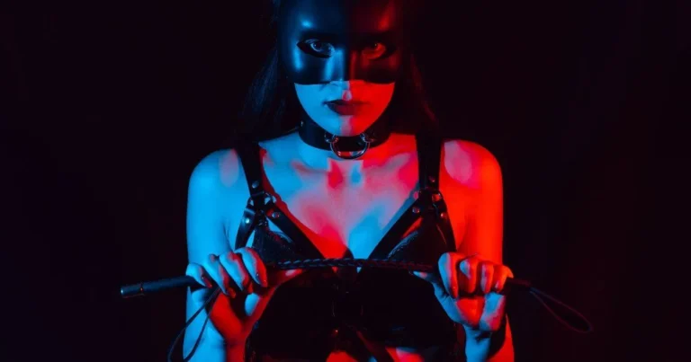 An image of a woman in lingerie with leather bondage straps. She is holding a riding crop, and starting BDSM play as she seductively looks into the camera.