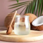 A jar of coconut oil against a pink background on a wooden turntable with coconuts behind the jar. This is the image for can you use coconut oil as lube.