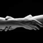Two hands holding each other at the forearm in a dark room rebuilding trust in BDSM.